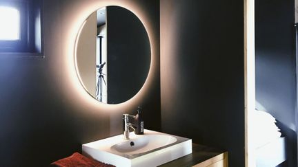 En hvit vask på en benk av lys eik i et mørkt og moderne bad med et speil som har baklys på seg