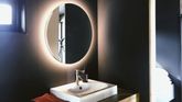 En hvit vask på en benk av lys eik i et mørkt og moderne bad med et speil som har baklys på seg