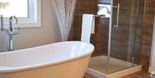 Et moderne bad med et hvitt badekar, en dusj med glassdører, en kontrastvegg av tre og et vindu over badekaret som slipper inn mye naturlig lys