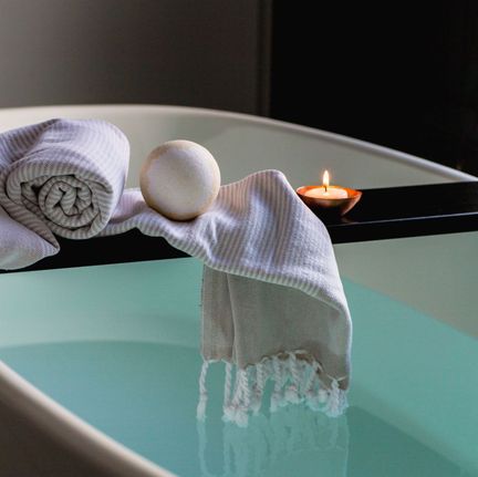 Et hvitt ovalt badekar som er fylt opp med vann og som har en treplanke over seg hvor det ligger et håndkle, en bathbomb og et tent telys i et i et rom med dempet belysning